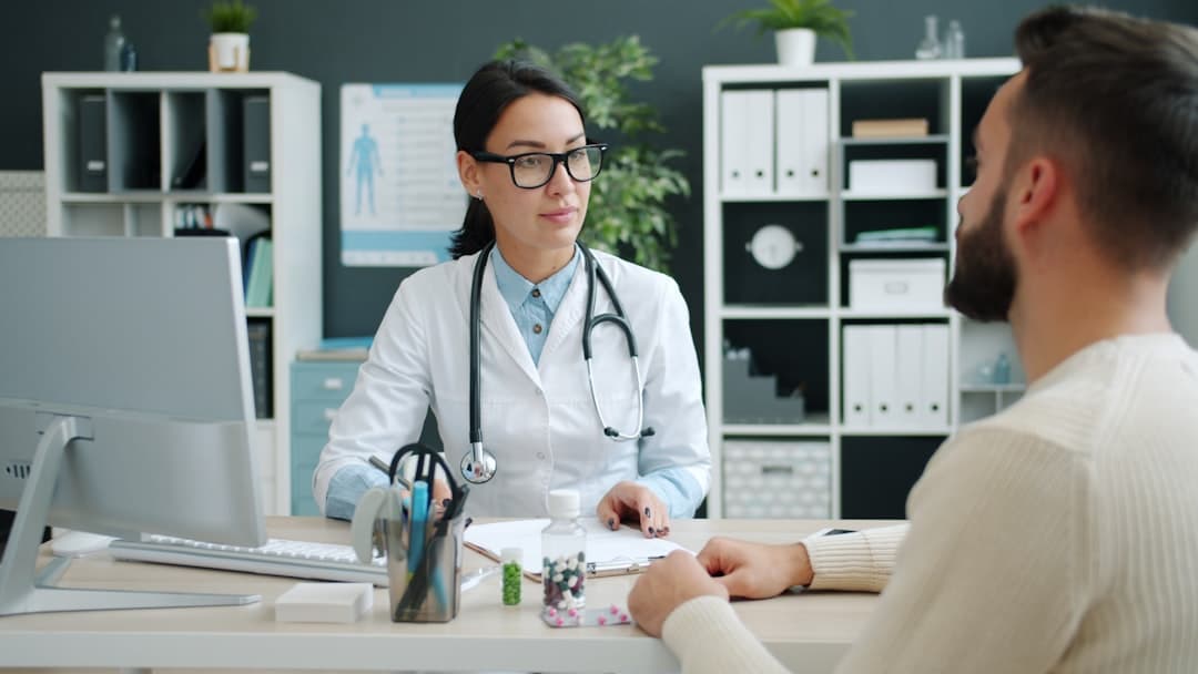 Friendly physician lady is talking to young man and filling in chart working in hospital room discussing disease symptoms and treatment. People and medicine concept.