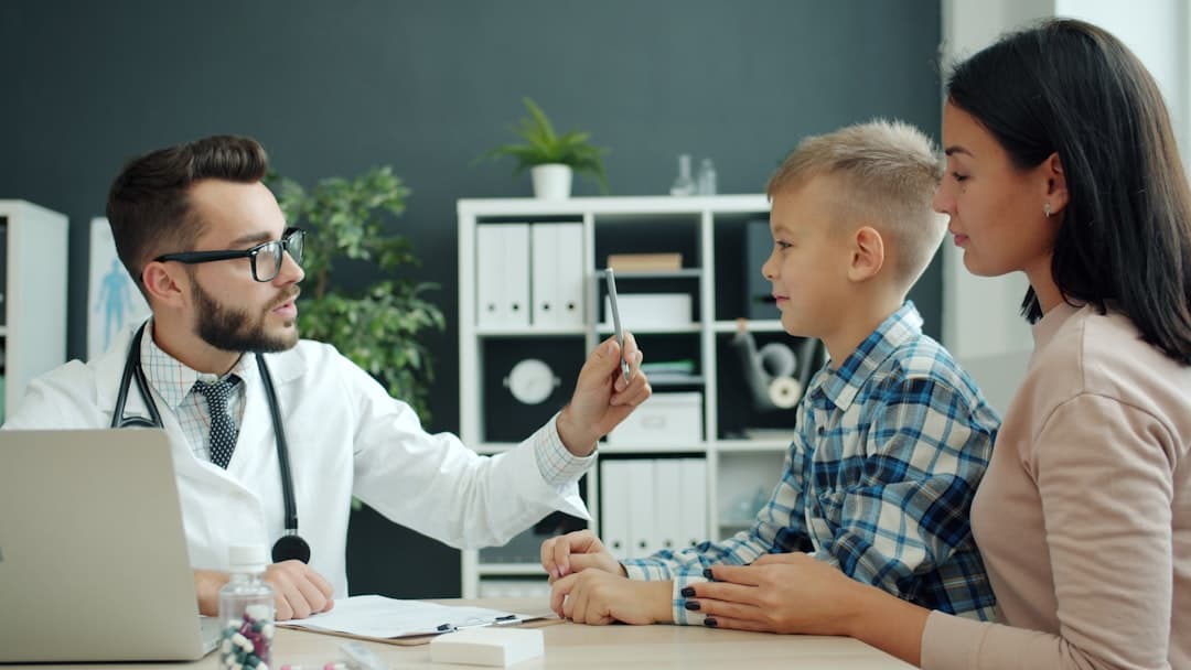 Male pediatrician in white gown is talking to family young mother and son examining child writing in chart during medical appointment in hospital.