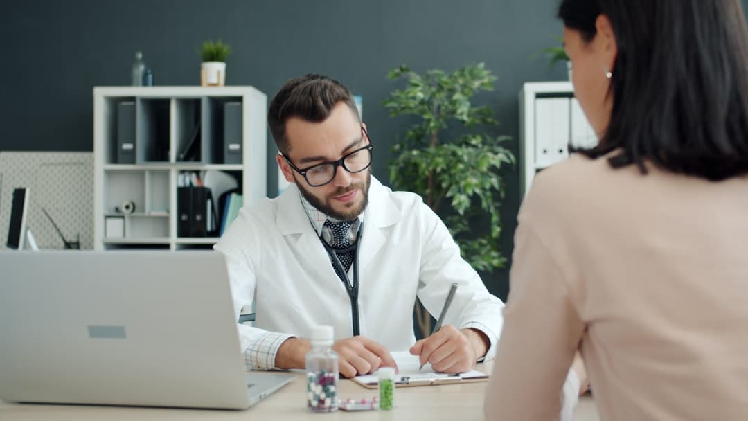 Young man doctor is consulting unhealthy female patient in clinic office talking writing in medical records sitting at table together. People and healthcare concept.